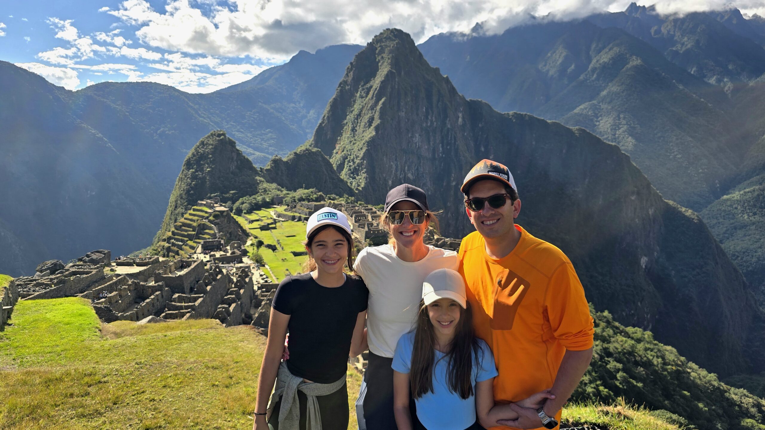 Una familia sonríe frente a Machu Picchu, con las terrazas de la ciudadela y el Huayna Picchu al fondo. El sol ilumina una de las Siete Maravillas del mundo moderno.