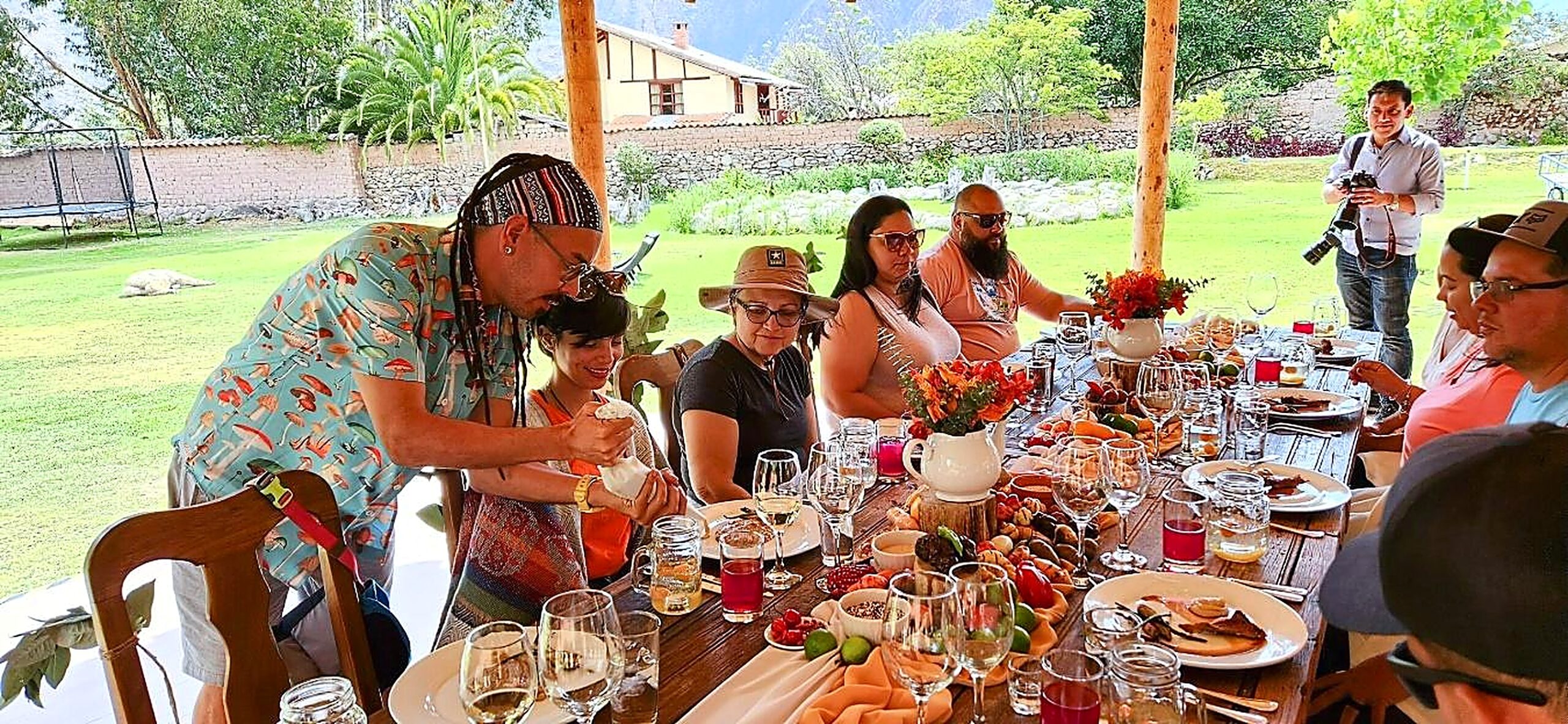 Viajeros comparten un almuerzo en una terraza de madera, con copas, flores, frutas y jarras sobre la mesa. Detrás quedan el jardín y una casa de campo.